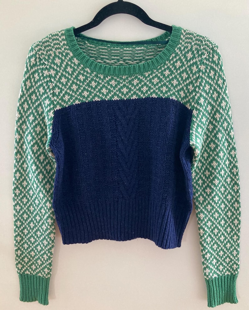 May include: A green and white patterned sweater with a blue cable knit panel in the center. The sweater has long sleeves and a round neckline.