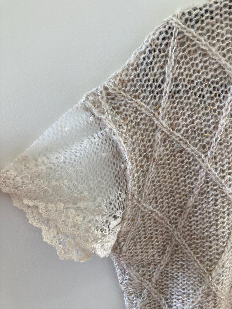 May include: A beige knitted sweater with a lace trim on the sleeves. The sweater has a cable knit pattern.