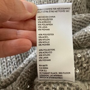 Op de afbeelding: Een wit label met zwarte tekst die de samenstelling van een kledingstuk beschrijft. Het label vermeldt "Hecho en China" (Gemaakt in China), "45% Polyester", "21% Acryl", "16% Polyamide", "12% Wol", "6% Mohair", "RN 66170", "CA 32054", "Voor EU", "24 Market Place, 3rd Floor", "Londen, VK WIWBAN", "Batch # 37W846X982C".