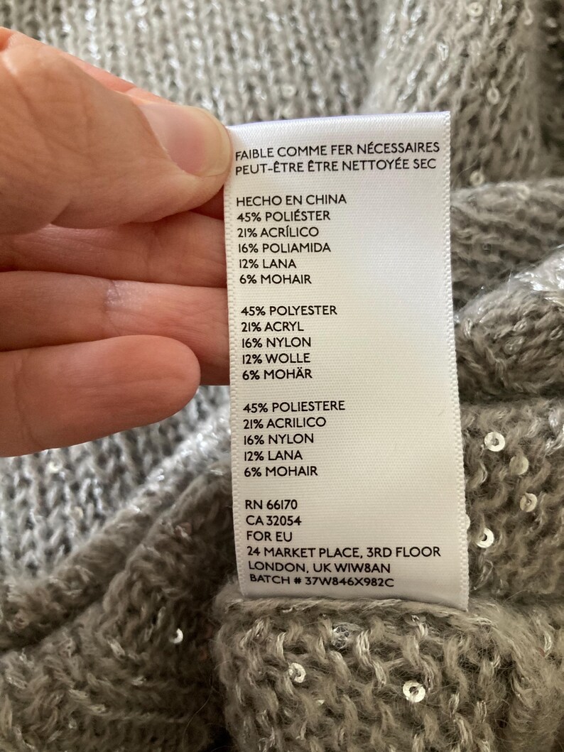 Op de afbeelding: Een wit label met zwarte tekst die de samenstelling van een kledingstuk beschrijft. Het label vermeldt "Hecho en China" (Gemaakt in China), "45% Polyester", "21% Acryl", "16% Polyamide", "12% Wol", "6% Mohair", "RN 66170", "CA 32054", "Voor EU", "24 Market Place, 3rd Floor", "Londen, VK WIWBAN", "Batch # 37W846X982C".