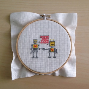 May include: Cross-stitch embroidery on white fabric in a wooden hoop, depicting two robots with red hearts and one robot holding a speech bubble with the text "00000000 00000000 00000000".