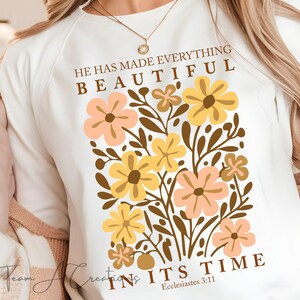 Puede incluir: Sudadera blanca con un dise&ntilde;o floral y el texto "&Eacute;l ha hecho todo hermoso en su tiempo, Eclesiast&eacute;s 3:11".