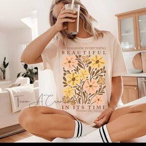 Puede incluir: Una camiseta beige con un dise&ntilde;o floral y el texto "&Eacute;l ha hecho todo hermoso en su tiempo, Eclesiast&eacute;s 3:11".