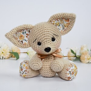 May include: A crocheted beige stuffed animal with floral fabric ears and feet. The animal has black eyes, a black nose, and a brown ribbon around its neck.