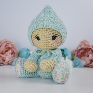 May include: A handmade crochet doll in a light blue outfit and matching pointed hat. The doll has a beige face with rosy cheeks and black eyes. The doll's feet are adorned with a floral fabric. The background features pink and blue flowers.