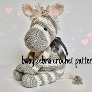 May include: A crocheted baby zebra toy with grey and white stripes. The zebra has a pink nose and a grey tail with a tassel. The toy is sitting on a white background with pink hearts.