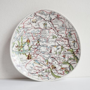 May include: A white ceramic plate featuring a detailed map design with red, green, and blue accents. The map includes place names, roads, and illustrations of people and animals. The plate has a slightly irregular shape.