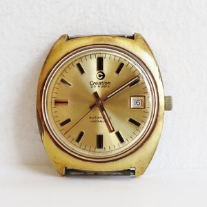 Puede incluir: Un reloj de pulsera dorado con una esfera redonda y agujas negras. La esfera del reloj tiene un fondo blanco con marcas negras y el texto "Creation 25 Rubis Automatic Incable" impreso en él. El reloj tiene una ventana de fecha en la posición de las 3 en punto.