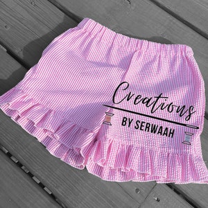 May include: Pink and white striped shorts with a ruffled hem. The shorts have a waistband and are made of a lightweight fabric. The text "Creations by Serwaah" is printed on the shorts.