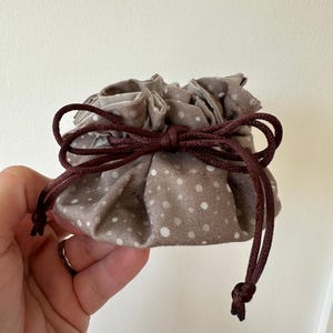 May include: A small, brown drawstring bag with white polka dots. The bag is made of fabric and has a brown drawstring closure.
