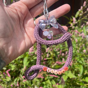 May include: A handmade keychain featuring a purple knitted letter "D" with the name "DELILA" spelled out in colourful beads. The keychain includes a clear bow and a silver chain, with a small charm. The background is a blurred view of green foliage.