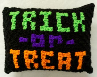 May include: A black, rectangular, crocheted pillow with the words "TRICK OR TREAT" in green, purple, and orange. The pillow is a Halloween-themed decoration.