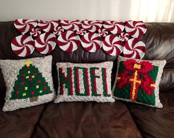 May include: Three crocheted throw pillows with Christmas themes. The pillows are on a brown leather couch with a white and red crocheted throw blanket behind them. The pillows feature a green Christmas tree, the word "NOEL", and a red and green gift box.