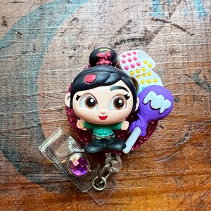 May include: A badge reel featuring a cartoon character with a black bun, green top, and black pants. The character is surrounded by a pink glitter heart, a number one, and a purple lollipop. The reel has a clear plastic holder with a purple gem.