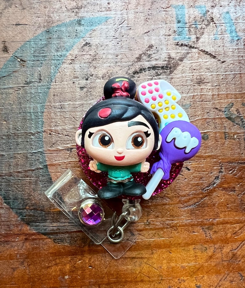 May include: A badge reel featuring a cartoon character with a black bun, green top, and black pants. The character is surrounded by a pink glitter heart, a number one, and a purple lollipop. The reel has a clear plastic holder with a purple gem.