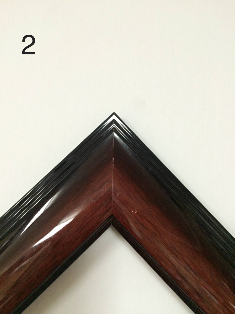 May include: A corner of a picture frame with a glossy, dark wood grain finish. The frame has a black outer edge and a number "2" in the upper left corner. The frame is angled, showcasing its polished surface.