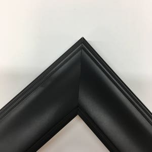 May include: A corner of a black picture frame. The frame has a matte finish and a beveled edge. The frame is made of wood and is designed to hold a photograph or artwork. The frame is against a white background.