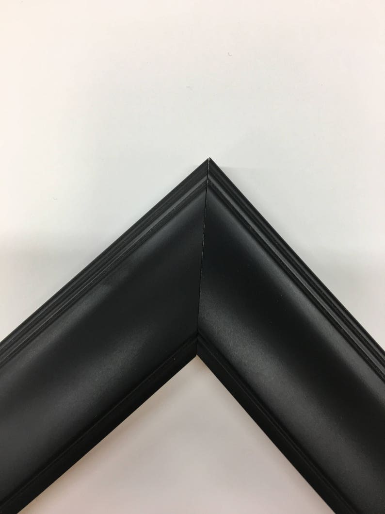May include: A corner of a black picture frame. The frame has a matte finish and a beveled edge. The frame is made of wood and is designed to hold a photograph or artwork. The frame is against a white background.