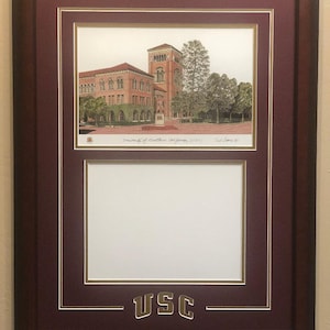 May include: A framed print of the University of Southern California (USC) campus. The print is in a maroon frame with a gold USC logo at the bottom. The print shows a building with a tower and trees in the foreground.