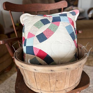 May include: A decorative pillow with a heart-shaped quilted design, featuring a patchwork of red, blue, and green fabrics. The pillow rests inside a rustic wooden basket, placed on a wooden chair.