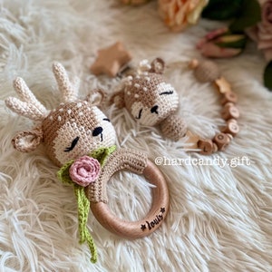 May include: A wooden baby teether with a crocheted deer head attached. The teether is engraved with the name "Louis" and has a pink flower on the deer's neck. The teether is on a white fluffy surface.