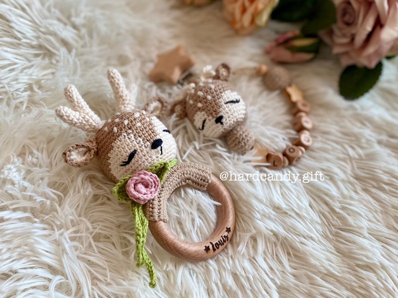 May include: A wooden baby teether with a crocheted deer head attached. The teether is engraved with the name "Louis" and has a pink flower on the deer's neck. The teether is on a white fluffy surface.
