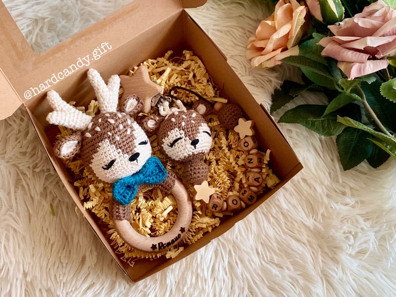 May include: A gift box filled with brown and white crocheted deer toys, a wooden star, and wooden letter blocks that spell "Romeo". The box is lined with brown shredded paper.