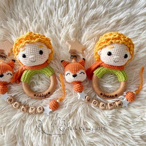 May include: Two handmade crochet baby toys. Each features a Little Prince doll with yellow hair, an orange scarf, and a green shirt. A wooden ring is attached, and a fox rattle hangs below. The names "A&euml;lys" and "Joann" are engraved on the rings.