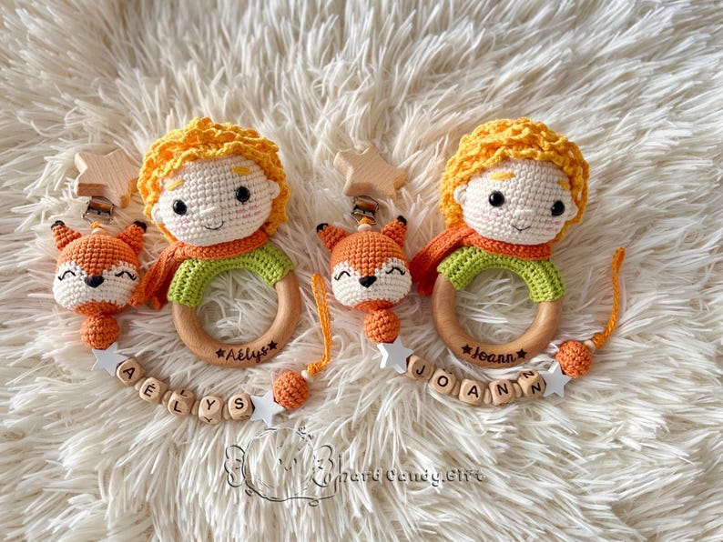 May include: Two handmade crochet baby toys. Each features a Little Prince doll with yellow hair, an orange scarf, and a green shirt. A wooden ring is attached, and a fox rattle hangs below. The names "A&euml;lys" and "Joann" are engraved on the rings.