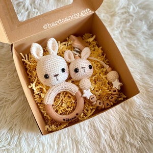 Peut inclure: Une boîte cadeau remplie de papier crépon jaune contient un hochet en forme de lapin crocheté avec un anneau en bois, un plus petit lapin crocheté attaché à un hochet en bois et une étoile, et un collier de perles en bois avec le nom "Leanne" écrit.