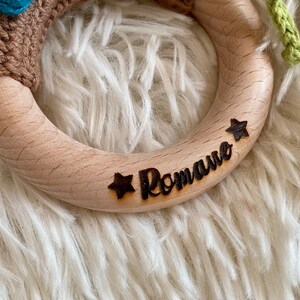 May include: A wooden ring with the name "Romane" and two stars engraved on it. The ring is light brown and has a smooth surface.