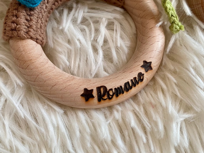 May include: A wooden ring with the name "Romane" and two stars engraved on it. The ring is light brown and has a smooth surface.