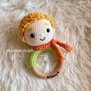 May include: A handmade crochet baby rattle featuring a smiling character with orange hair and scarf, and a green shirt. The rattle has a wooden ring base with the name "Noha" and the text "hardcandy.gift".