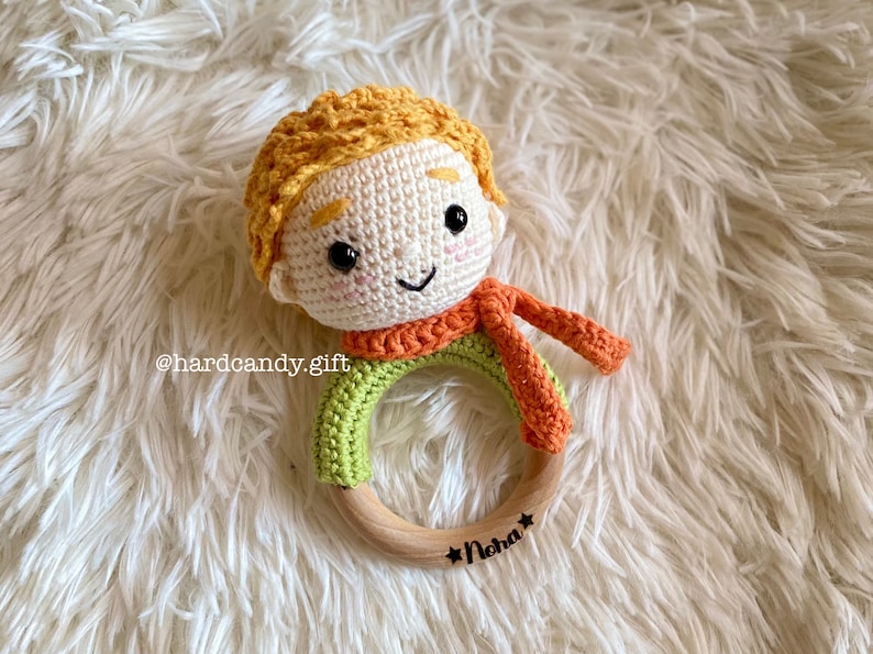 May include: A handmade crochet baby rattle featuring a smiling character with orange hair and scarf, and a green shirt. The rattle has a wooden ring base with the name "Noha" and the text "hardcandy.gift".
