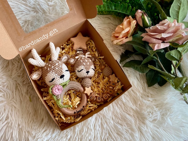 May include: A gift box filled with brown shredded paper and two crocheted deer toys. One deer is attached to a wooden teething ring with the name "Louis" engraved on it. The other deer is attached to a wooden star-shaped teether. The box also contains a wooden bead and a set of wooden letter beads that spell out "Louis".