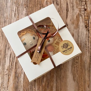 May include: A white gift box with a clear window, tied with a brown ribbon, contains two crocheted dolls with orange hair. The box has a sticker that says "Hard Candy make it sweeter". The dolls are on a wooden surface.