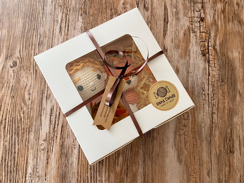 May include: A white gift box with a clear window, tied with a brown ribbon, contains two crocheted dolls with orange hair. The box has a sticker that says "Hard Candy make it sweeter". The dolls are on a wooden surface.