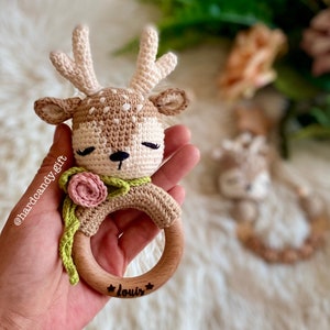 May include: A crocheted deer rattle with a pink flower and green leaves, attached to a wooden ring with the name "Louis" engraved on it.