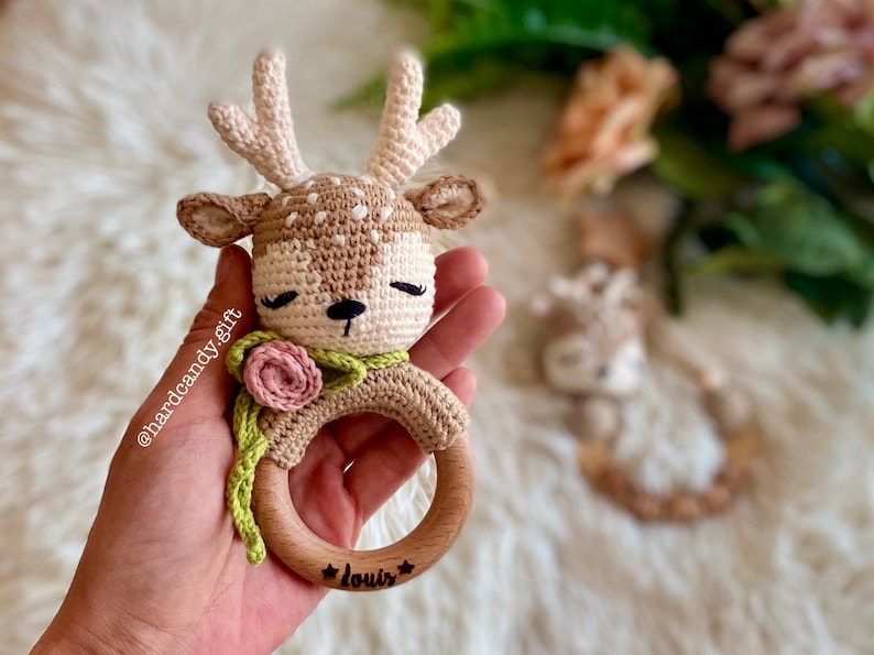 May include: A crocheted deer rattle with a pink flower and green leaves, attached to a wooden ring with the name "Louis" engraved on it.