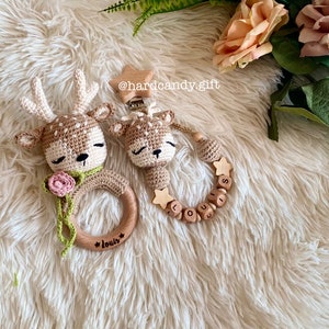 May include: Two crocheted deer toys, one with a wooden ring and the other with a wooden bead necklace. The ring has the name "Louis" engraved on it and the necklace has the name "Louis" spelled out with wooden beads. The toys are on a white fluffy surface.
