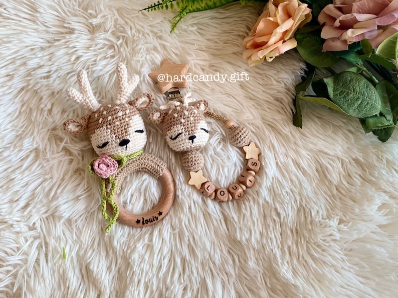 May include: Two crocheted deer toys, one with a wooden ring and the other with a wooden bead necklace. The ring has the name "Louis" engraved on it and the necklace has the name "Louis" spelled out with wooden beads. The toys are on a white fluffy surface.