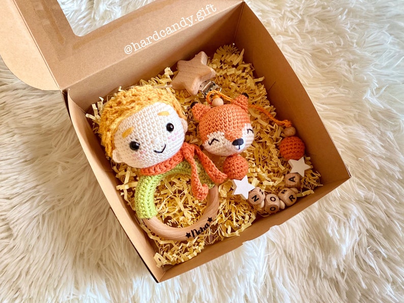 May include: A gift box containing a crocheted Little Prince doll with a wooden ring teether, a crocheted fox toy, and a pacifier clip with wooden beads. The box is filled with yellow paper shreds and includes wooden star accents.