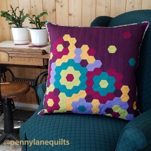 Puede incluir: Una almohada acolchada colorida con un patr&oacute;n geom&eacute;trico de hex&aacute;gonos en tonos de morado, turquesa, amarillo y rojo. La almohada est&aacute; sentada en un sill&oacute;n verde oscuro con una tela acolchada.