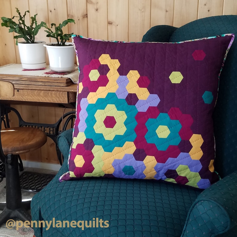 Puede incluir: Una almohada acolchada colorida con un patr&oacute;n geom&eacute;trico de hex&aacute;gonos en tonos de morado, turquesa, amarillo y rojo. La almohada est&aacute; sentada en un sill&oacute;n verde oscuro con una tela acolchada.