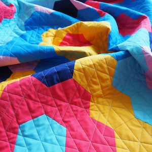 Puede incluir: Una colcha colorida con un patr&oacute;n geom&eacute;trico en tonos de azul, rosa, amarillo y rojo. La colcha est&aacute; hecha de cuadrados de tela cosidos juntos para crear un patr&oacute;n hexagonal. La colcha est&aacute; doblada y drapeada sobre una superficie.