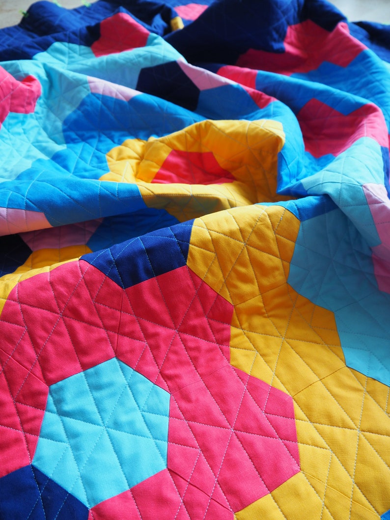Puede incluir: Una colcha colorida con un patr&oacute;n geom&eacute;trico en tonos de azul, rosa, amarillo y rojo. La colcha est&aacute; hecha de cuadrados de tela cosidos juntos para crear un patr&oacute;n hexagonal. La colcha est&aacute; doblada y drapeada sobre una superficie.