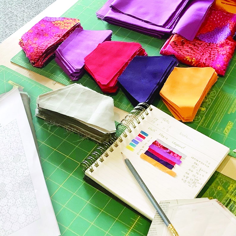 Puede incluir: Una estera de corte verde con recortes de tela en varios colores y patrones, incluyendo rosa, morado, rojo, naranja y blanco. Un cuaderno con notas escritas a mano y muestras de tela est&aacute; sobre la estera. El cuaderno tiene las palabras "Mitad", "Entero", "Repetir" y "Total" escritas en &eacute;l.