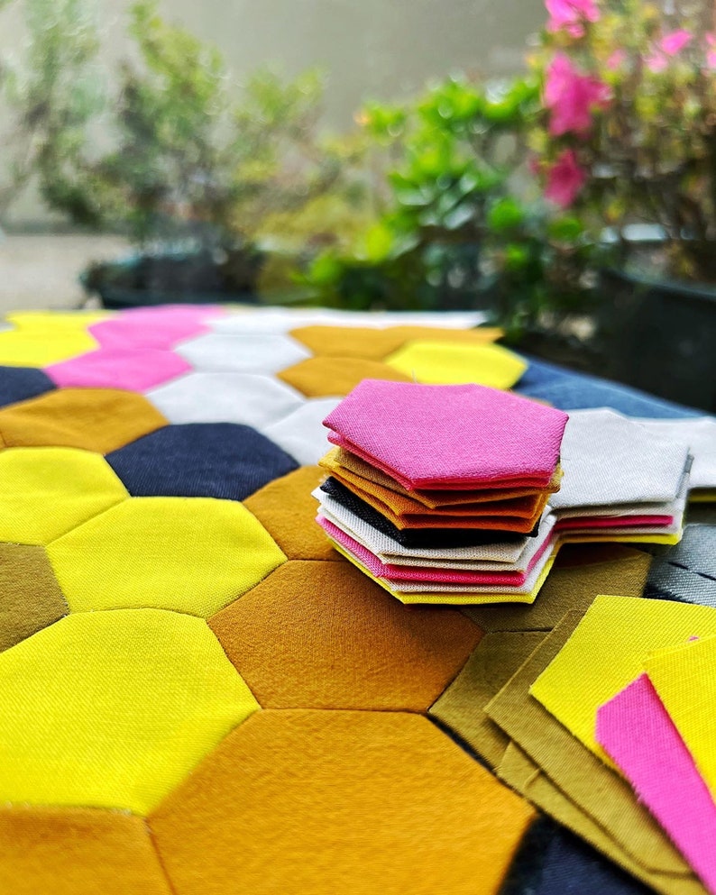 Puede incluir: Una pila de piezas de tela hexagonales en varios colores, incluyendo amarillo, naranja, rosa, blanco y negro. Las piezas de tela est&aacute;n dispuestas en un patr&oacute;n de panal en una mesa.
