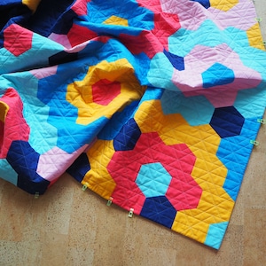 Puede incluir: Una colcha colorida con un patr&oacute;n geom&eacute;trico en tonos de azul, rosa, amarillo y rojo. La colcha est&aacute; doblada y sujeta a un suelo de madera.