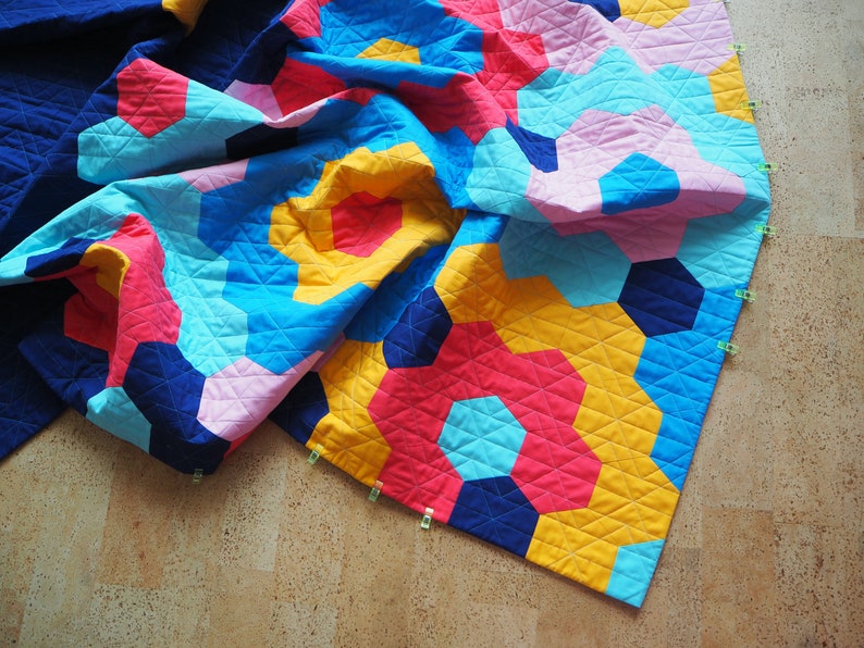 Puede incluir: Una colcha colorida con un patr&oacute;n geom&eacute;trico en tonos de azul, rosa, amarillo y rojo. La colcha est&aacute; doblada y sujeta a un suelo de madera.